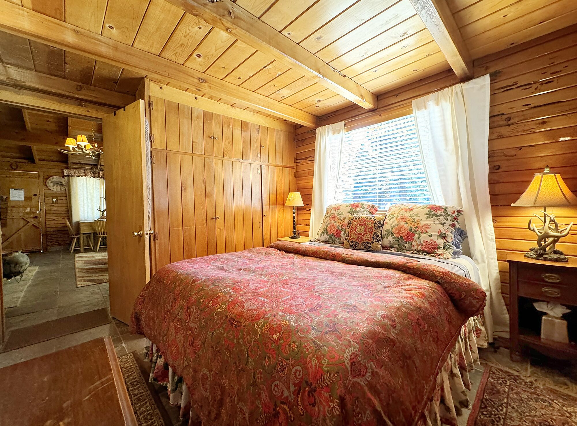 Ein Schlafzimmer einer Holzhütte mit einem großen Bett, einem Fenster mit Vorhängen, einer Holzbalkendecke und einem kleinen Tisch mit einer Lampe.