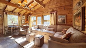 A cozy log cabin interior with a wooden ceiling, a sofa, a table with chairs, and a window with curtains.