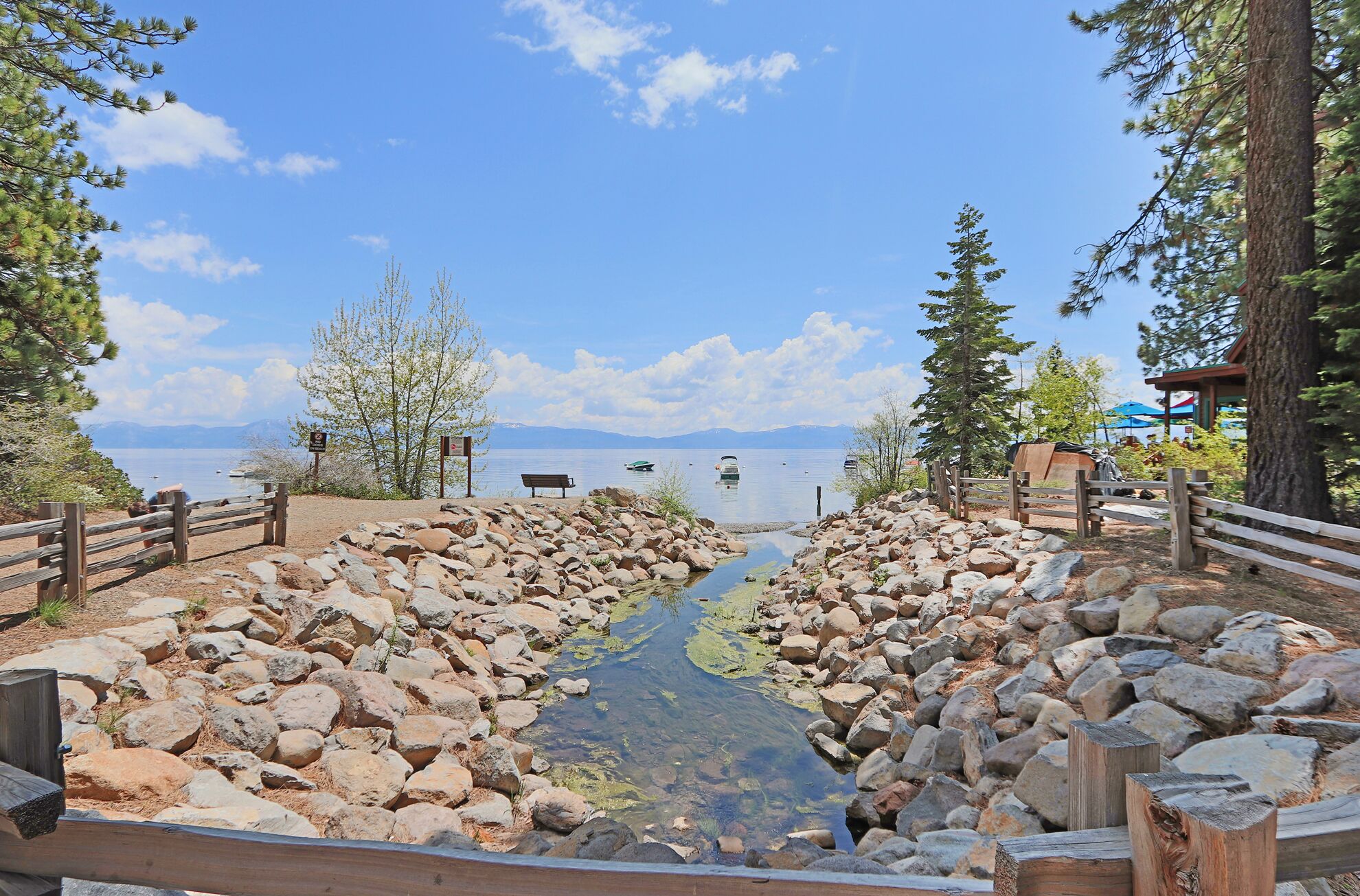 A rocky shoreline with a wooden walkway leading to a lake, surrounded by trees and a distant mountain range.