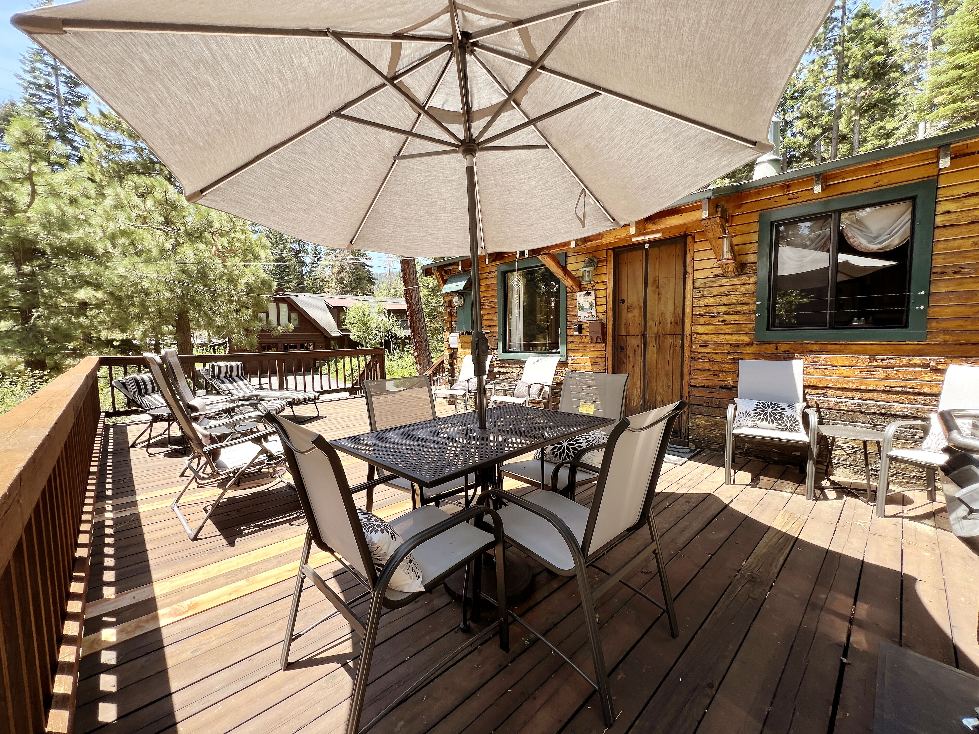Una terraza de madera con una mesa y sillas, un paraguas grande y una cabaña al fondo.