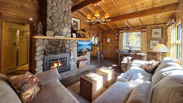 A cozy log cabin interior with a stone fireplace, wooden ceiling beams, a comfortable sofa, and a television.