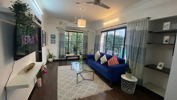 Ein modernes Wohnzimmer mit einem blauen Sofa, einem Glastisch und einem Teppich mit weißem Muster. Es gibt einen Deckenventilator, einen an der Wand montierten Fernseher und ein großes Fenster mit Vorhängen.