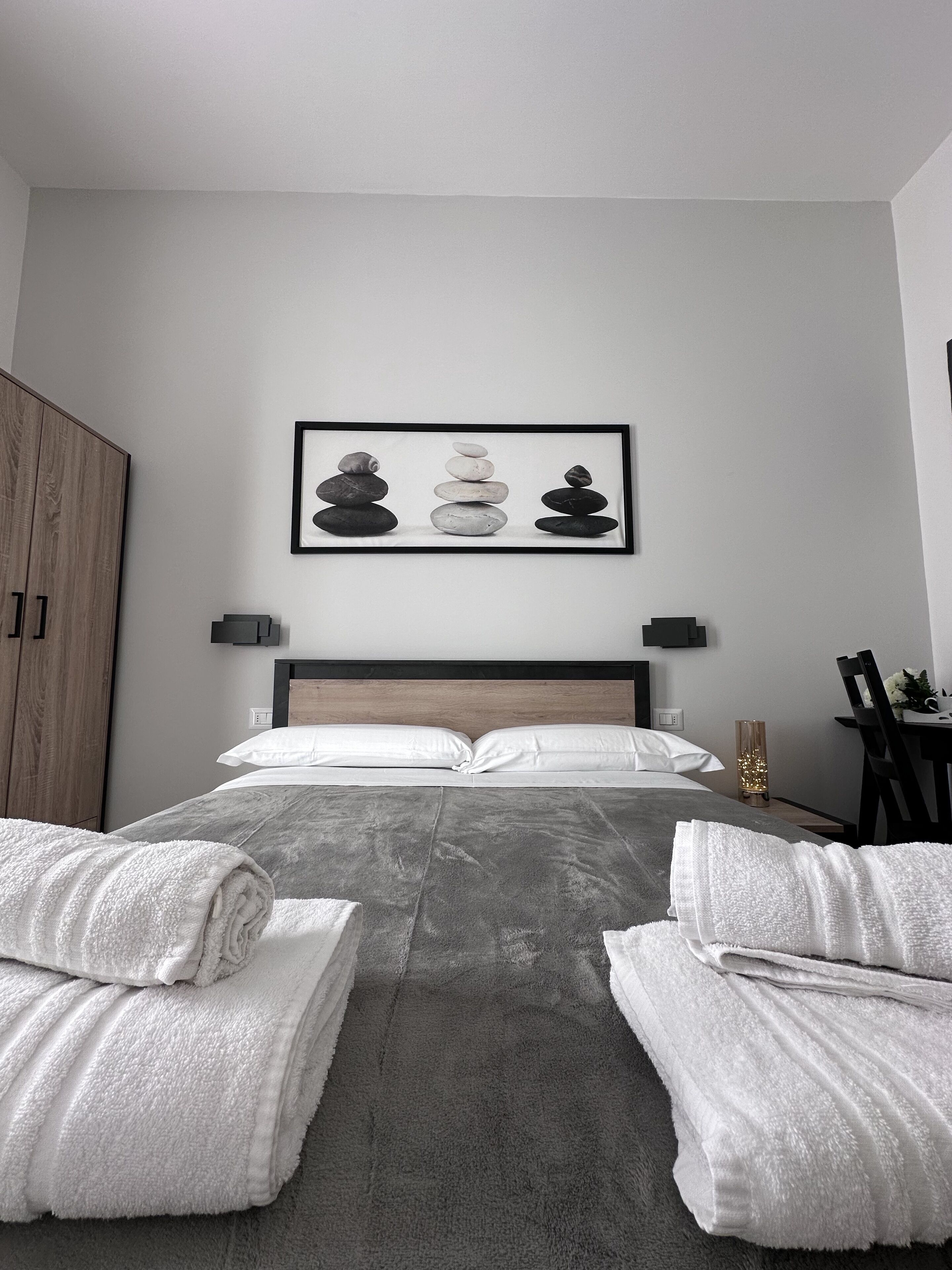 A neatly made bed with white pillows and towels, a framed artwork of stacked stones on the wall, and a wooden wardrobe to the left.