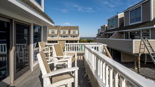 A deck with white railings and chairs, overlooking a residential area with beige houses and a clear sky.