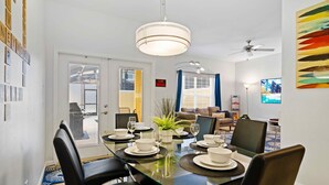 A dining area with a glass table set for four, surrounded by black chairs. There is a ceiling fan, a potted plant, and a television on the wall.