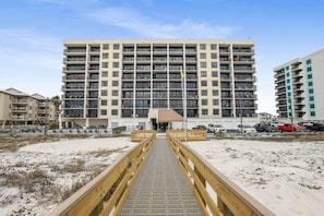 Ein mehrstöckiges Hotel mit zentralem Eingang, ein Sandstrand im Vordergrund und ein hölzerner Steg, der zum Eingang führt.