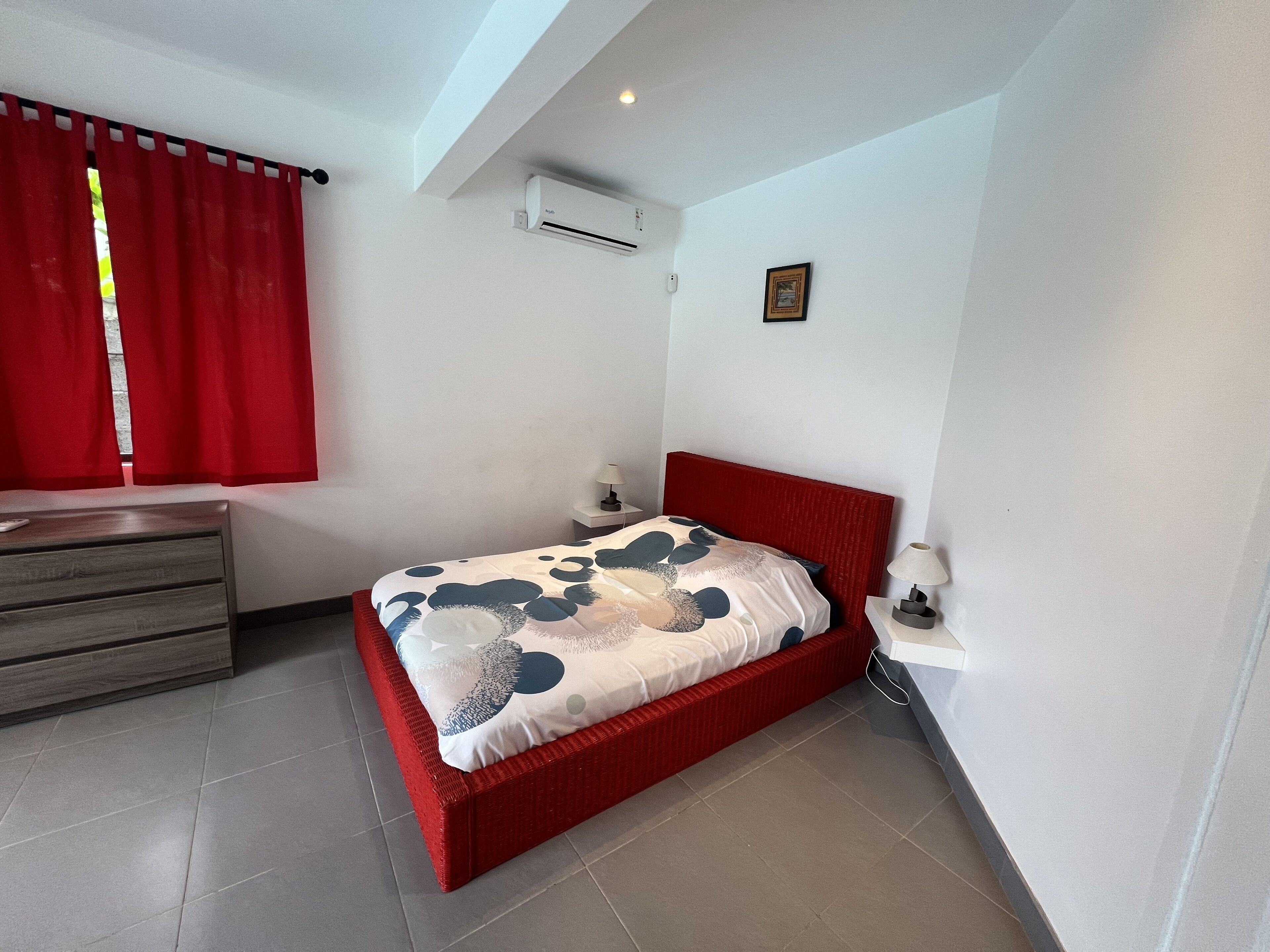 A bedroom with a red bed, a wooden dresser, a red curtain, an air conditioning unit, and a small picture on the wall.