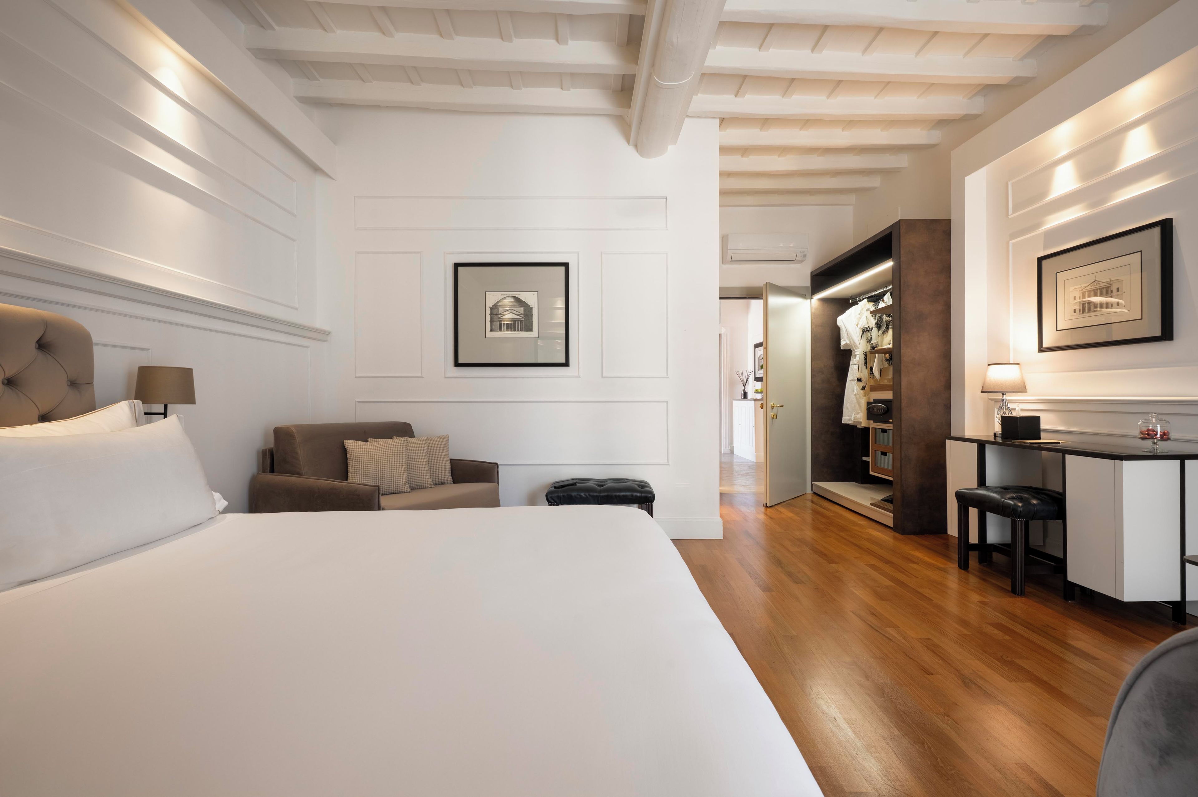 A hotel room with a large bed, a sofa, a desk, and a wardrobe. There are framed pictures on the walls and a ceiling with exposed beams.