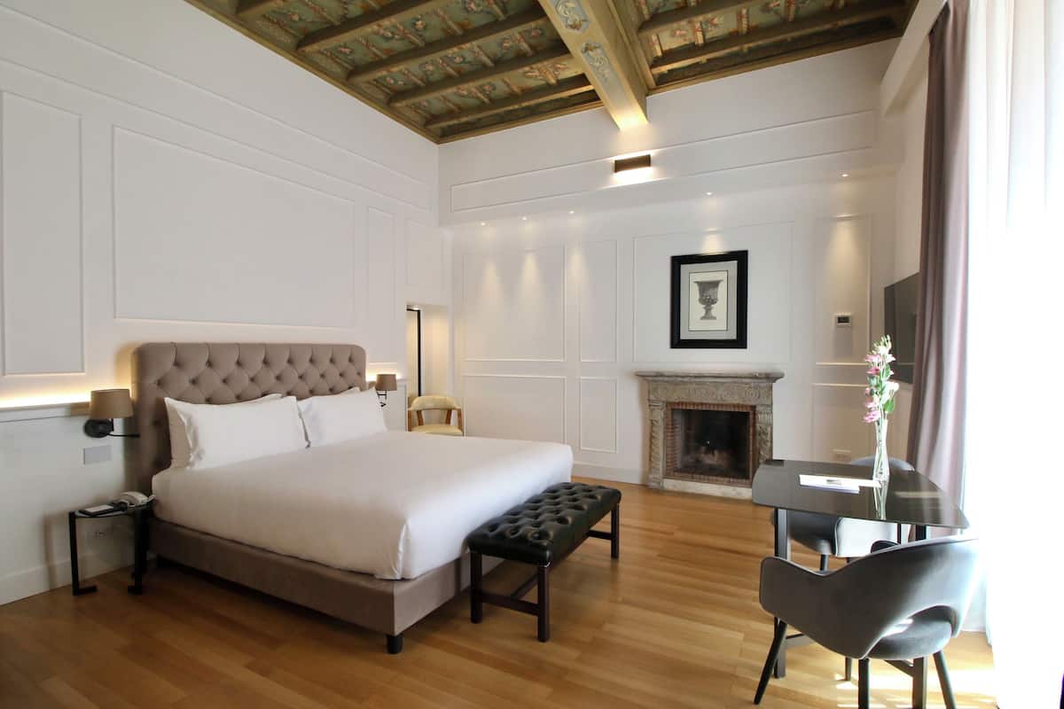 A bedroom with a large bed, a bedside table, a chair, a fireplace, and a TV.