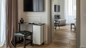 A modern hotel room with a flat-screen TV mounted on the wall, a desk with a chair, a large bathtub, and a wooden floor. - Palazzo Goldoni Condotti Collection (Rome)