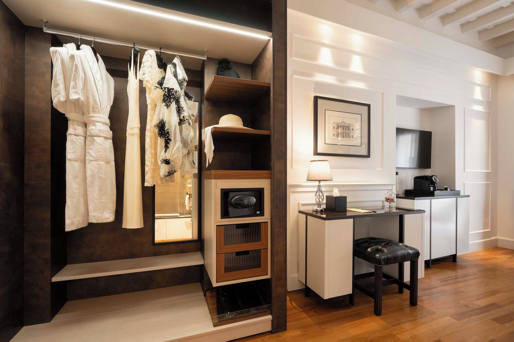 A modern hotel room with a walk-in closet, a desk with a lamp, and a flat-screen TV.