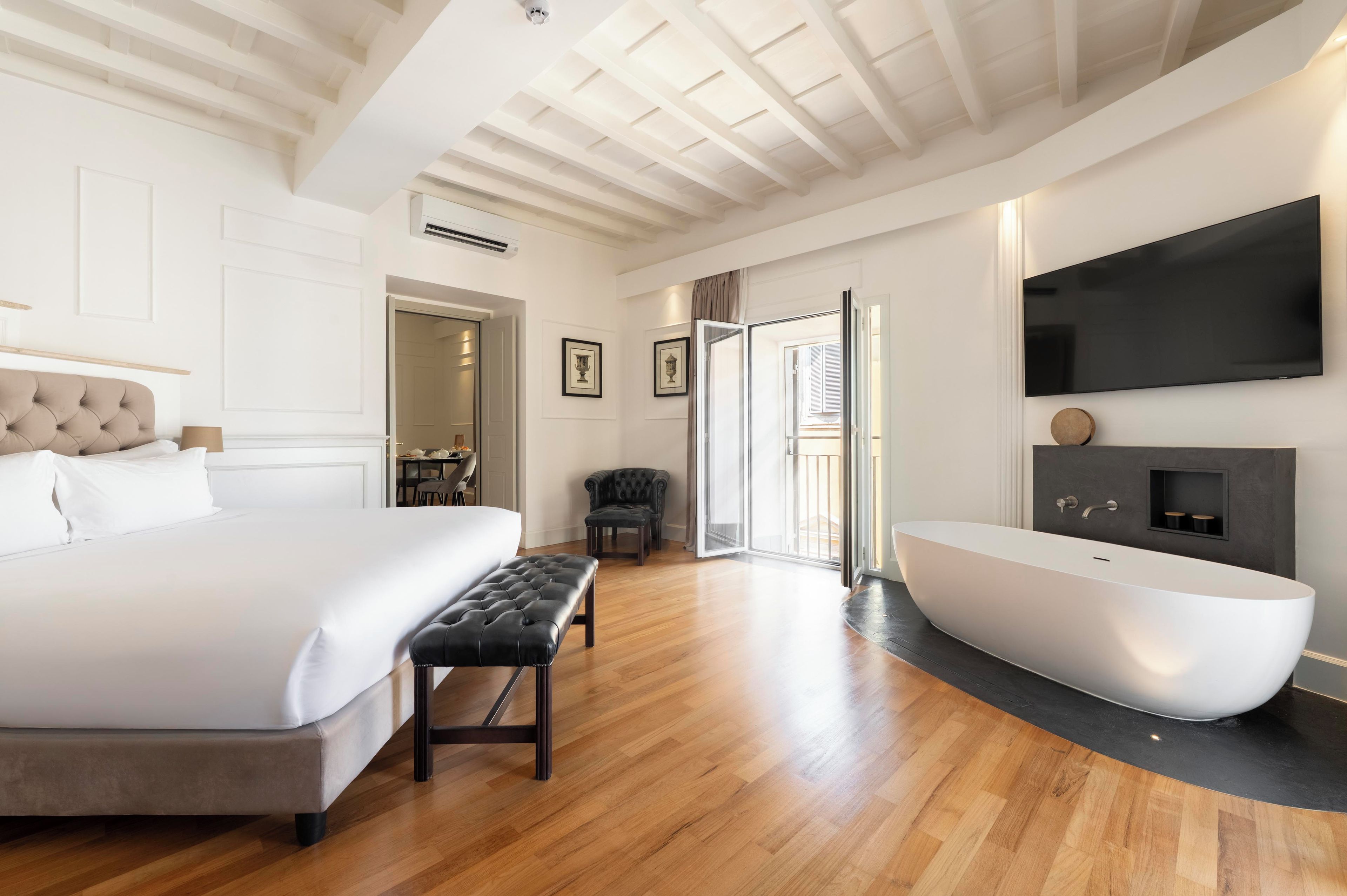 A modern bedroom with a large bed, a bench, a flat-screen TV, and a freestanding bathtub.