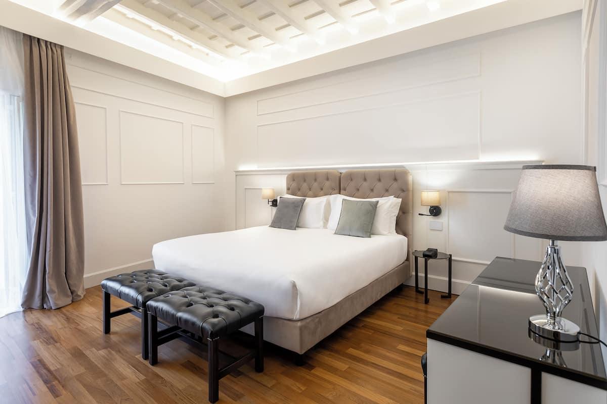 A modern hotel room with a large bed, a bench, a desk, and a lamp.