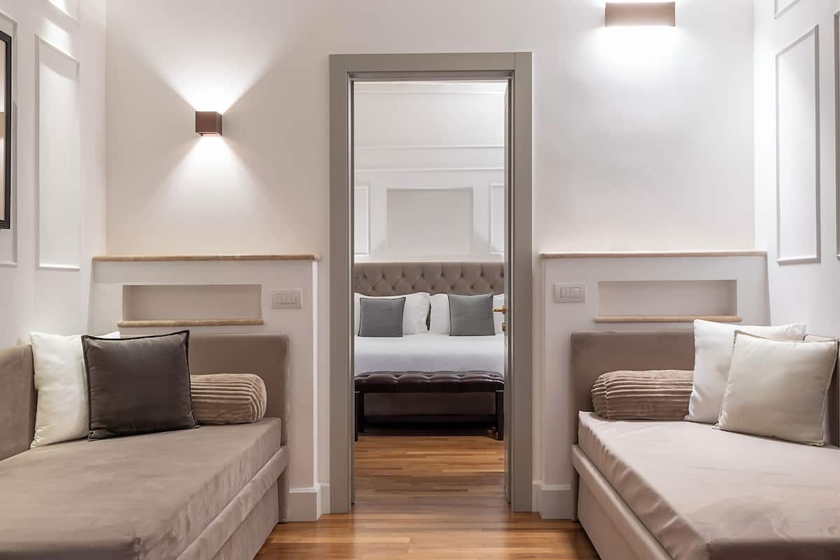 A modern hotel room with a wooden floor, two sofas, a bed with a headboard, and a mirror on the wall.