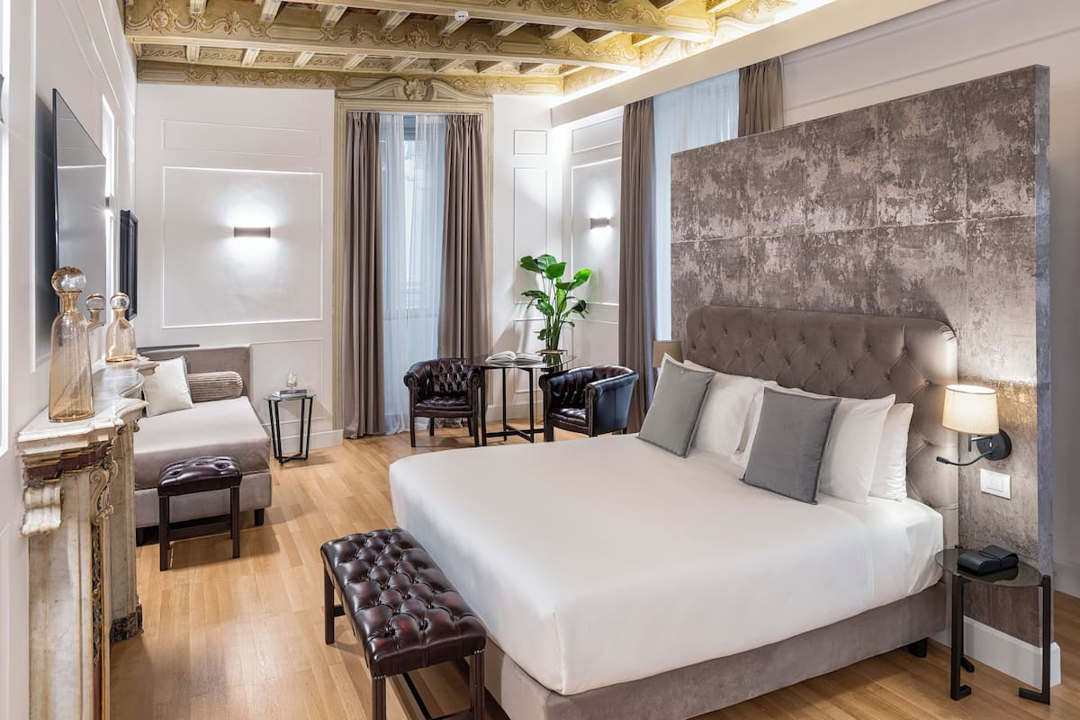 A modern hotel room with a large bed, a seating area with a bench and chairs, a small table, and a decorative plant.