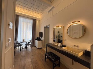A hotel room with a wooden floor, a dining area with a table and chairs, a large mirror, and a modern bathroom with a sink and vanity. - Palazzo Goldoni Condotti Collection (Rome)