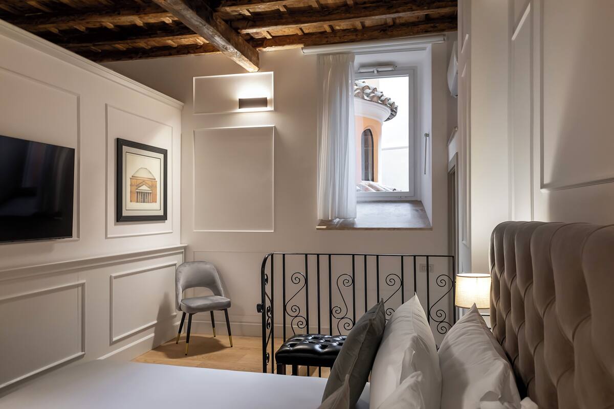 A bedroom with a bed, a chair, a TV, a window with a view of a building, and a lamp.