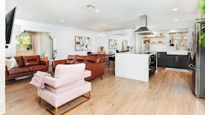 A modern living room with a brown leather sofa, a pink armchair, and a dining area with a table and chairs. A kitchen with a white countertop and a refrigerator is visible in the background. - Boca Family Fun & Charm - Villa Near Downtown & Beach (Boca Raton)
