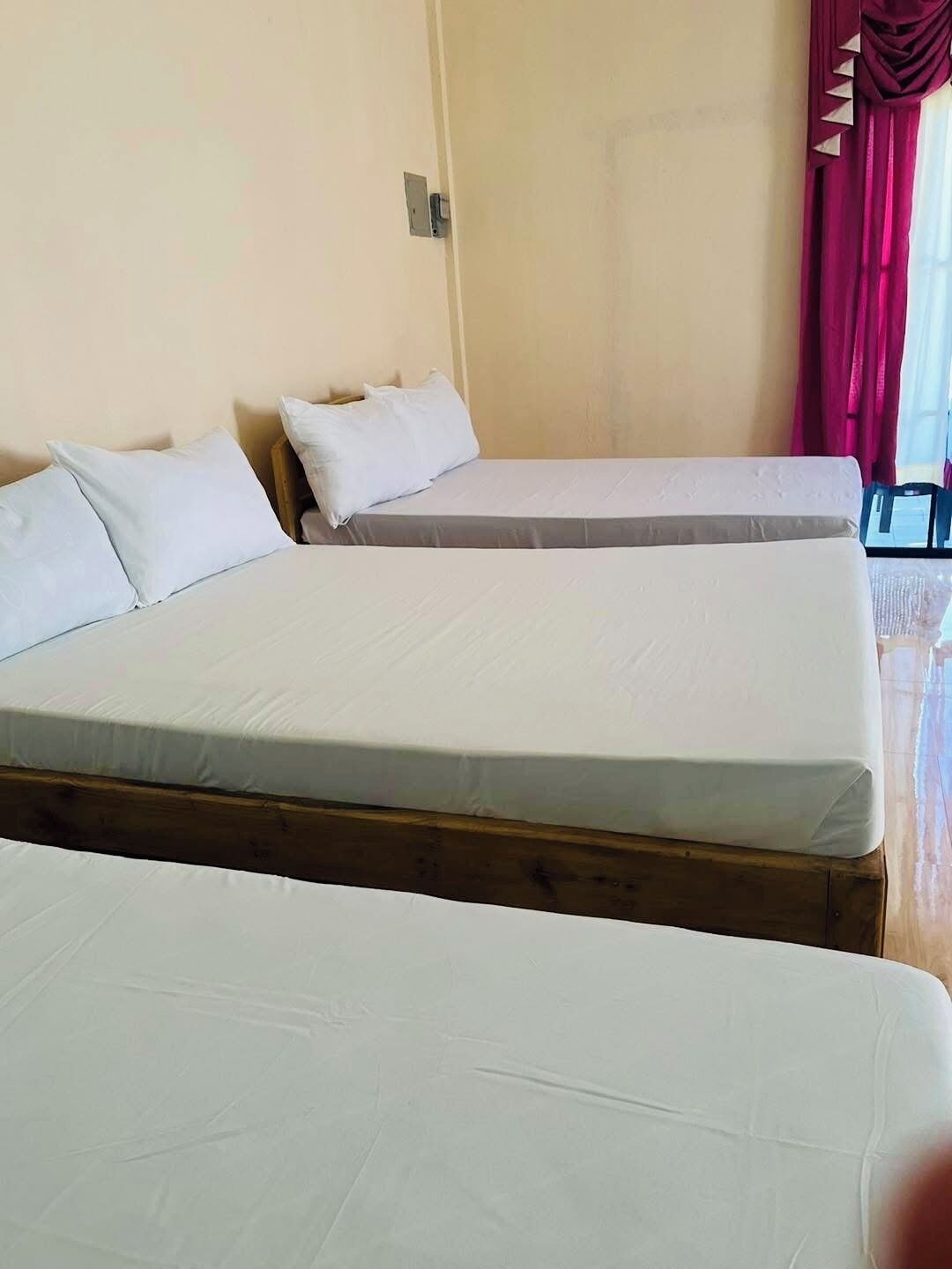 A room with two single beds, a wooden headboard, a wall socket, and a window with red curtains.