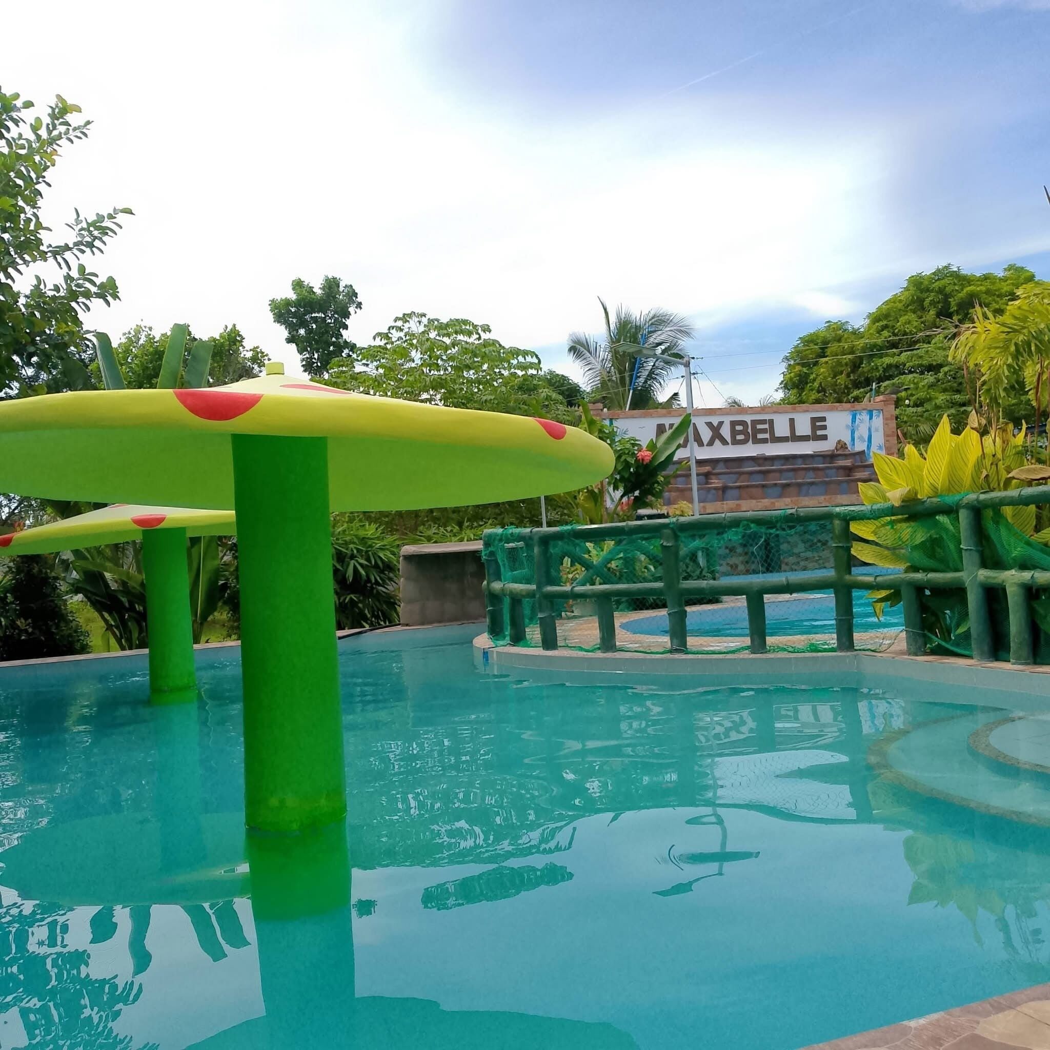 Una piscina con una estructura verde y amarilla en primer plano, rodeada de exuberante follaje y un letrero que dice 'MAXBELLE'.