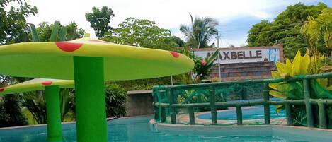Una piscina con una estructura verde y amarilla en primer plano, rodeada de exuberante follaje y un letrero que dice 'MAXBELLE'.