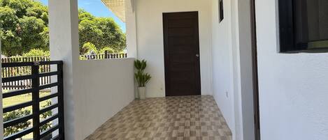 A covered corridor with a checkered floor, a potted plant, and a door at the end.