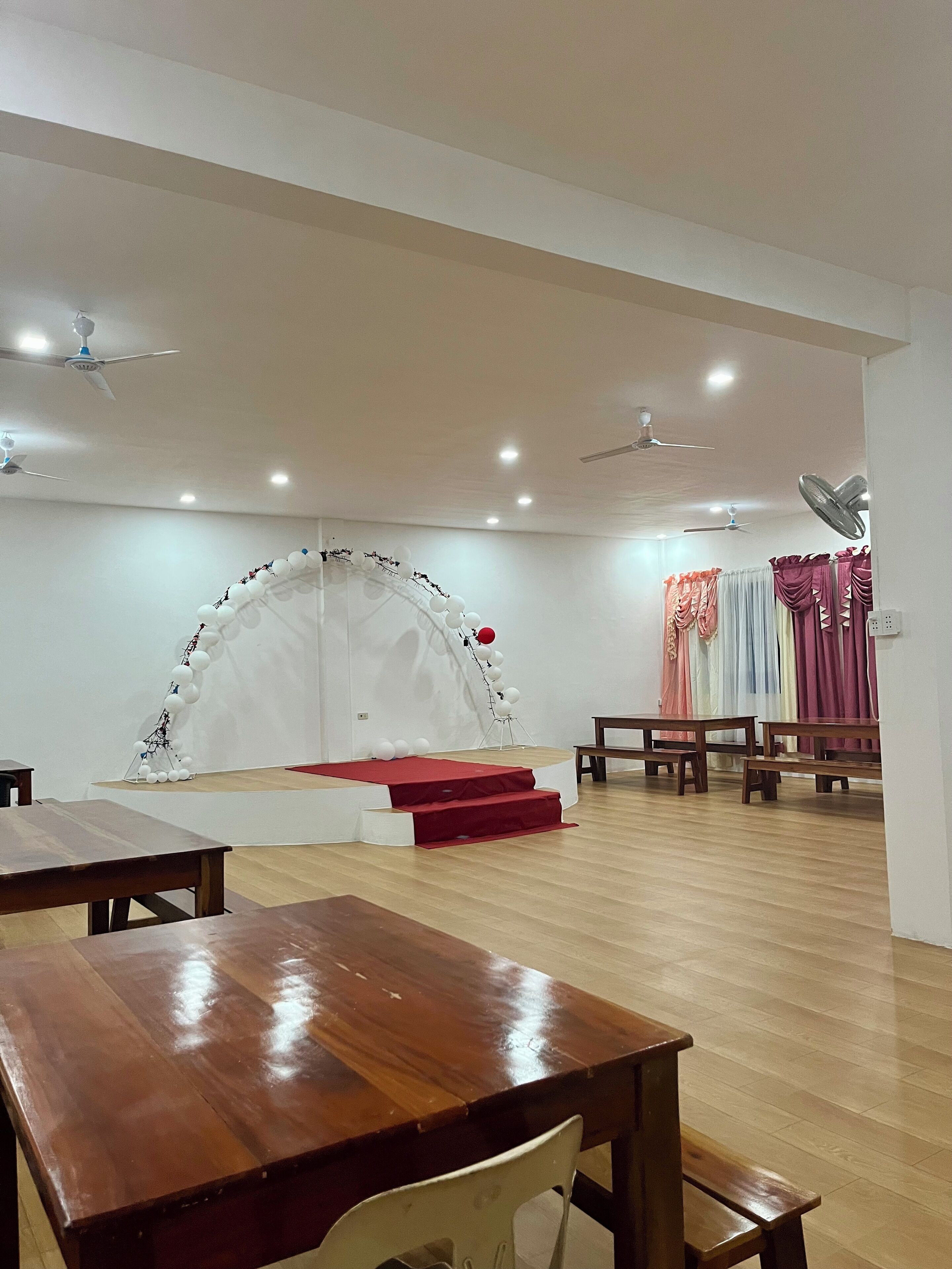A spacious room with a stage, wooden tables, and ceiling fans.