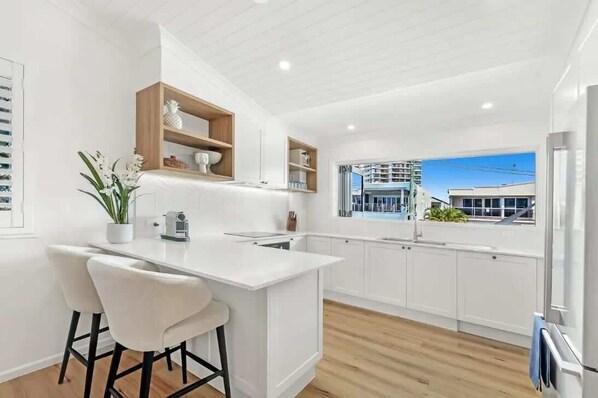 A modern kitchen with white cabinetry, a central island with bar stools, a wooden shelf above the island, and a view of a high-rise building through a window.
