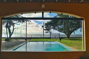 Vista a través de un amplio ventanal de una piscina con vista despejada al mar y un pabellón.