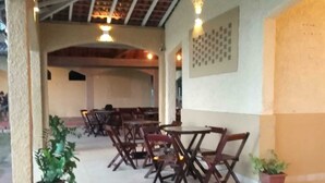 Un patio al aire libre con mesas y sillas de madera, plantas en macetas y un techo con vigas a la vista.