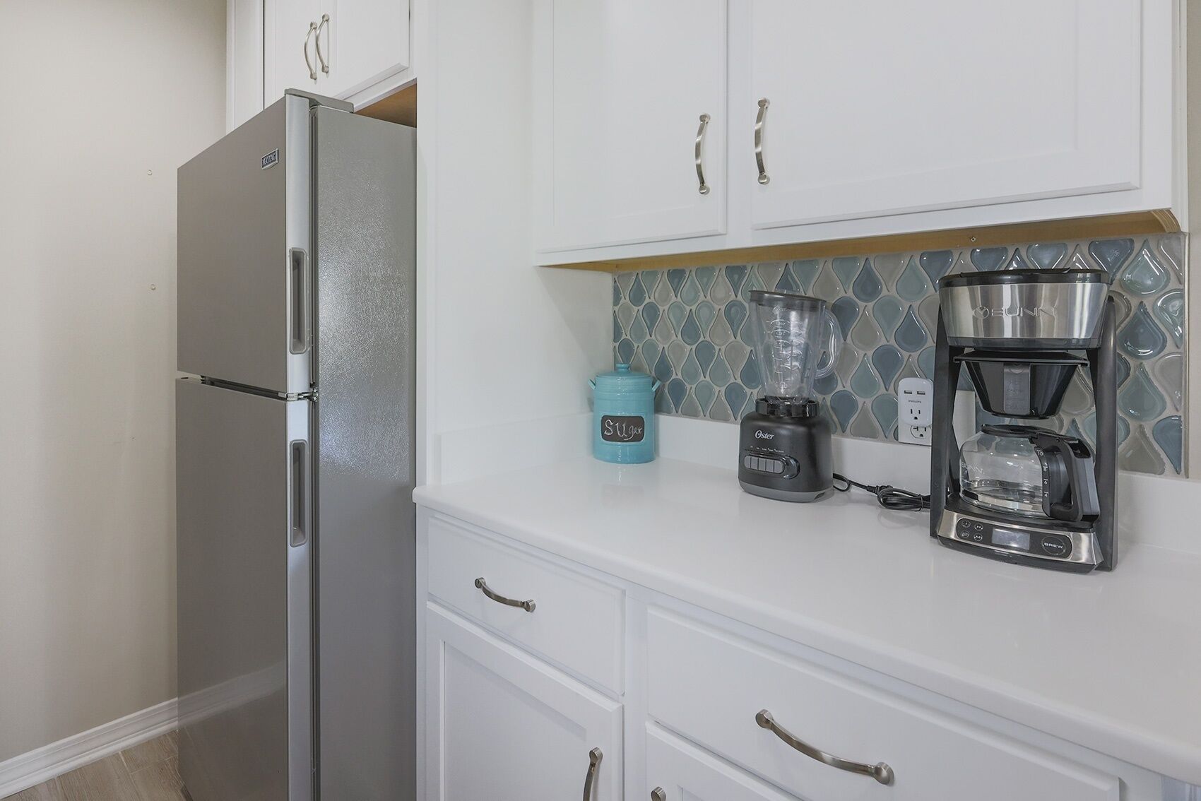 Una cocina con gabinetes blancos, un refrigerador de acero inoxidable, una licuadora y una cafetera.