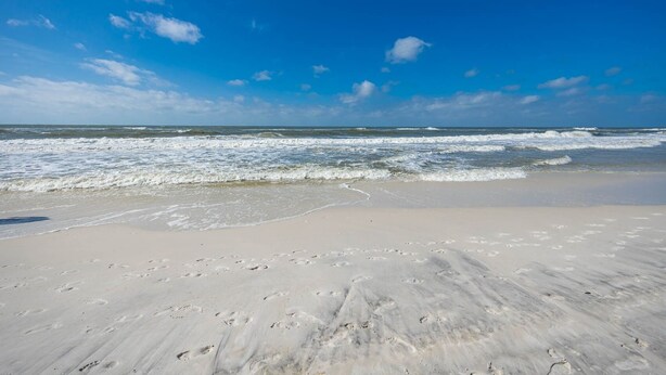 A beach with footprints leading to the water, a clear sky, and waves approaching the shore.