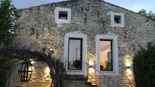 Un bâtiment en pierre avec deux fenêtres et une porte, un escalier menant à l’entrée et des sièges à l’extérieur.