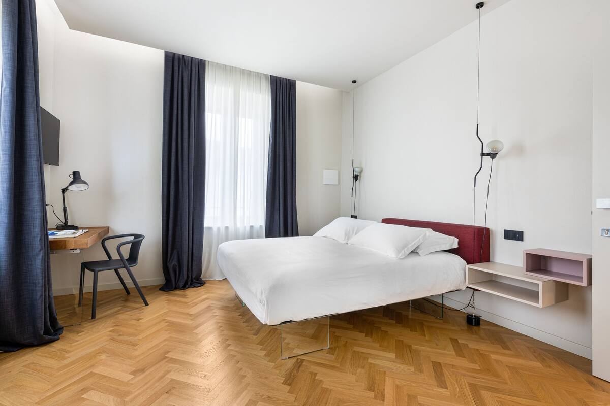 Quarto de hotel moderno com piso de madeira em padrão herringbone, cama com lençóis brancos, escrivaninha com cadeira e televisão fixada na parede.