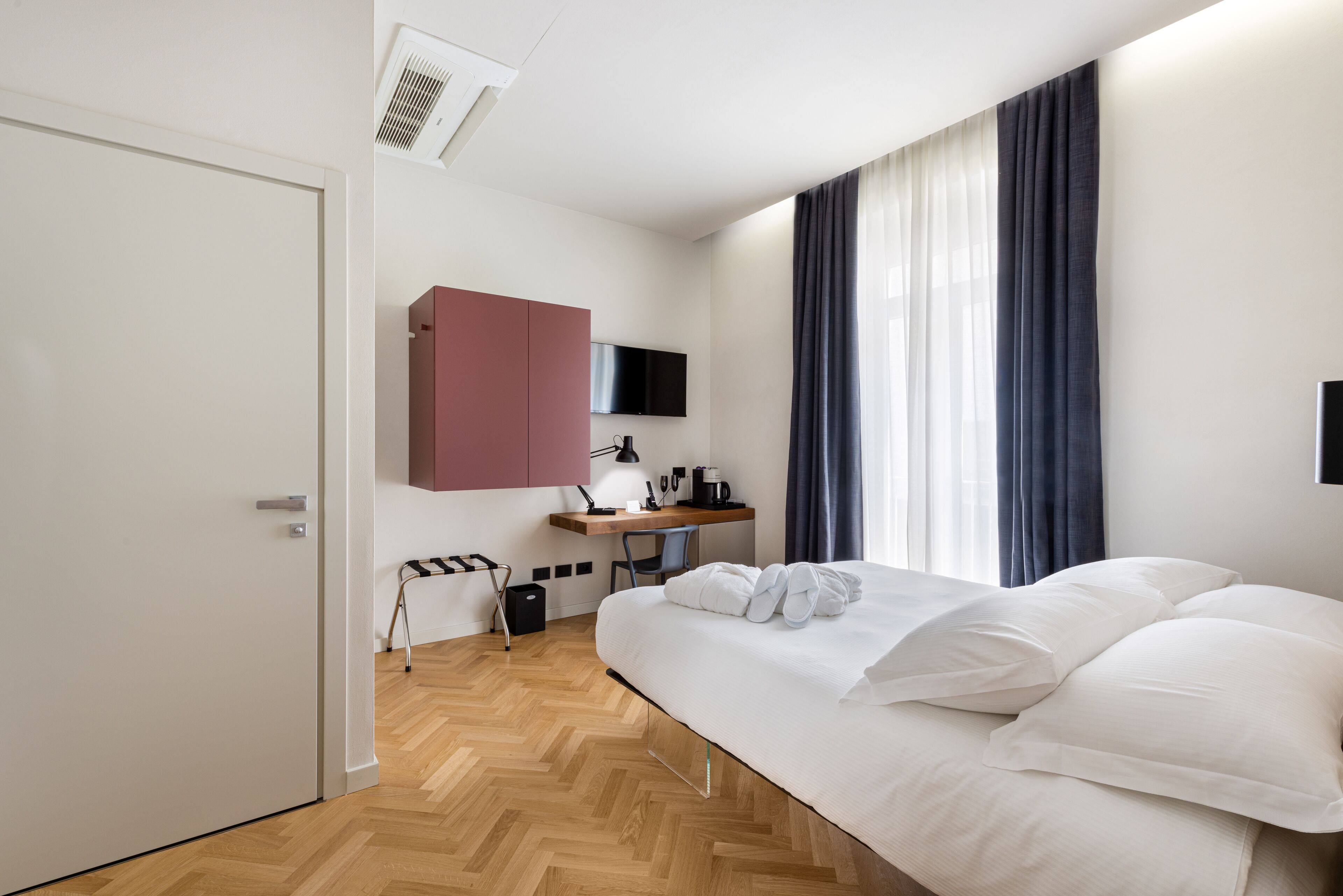 Una camera d'albergo moderna con pavimento in legno a spina di pesce, un ampio letto con lenzuola bianche, una scrivania con un computer e una televisione a schermo piatto montata a parete, e una finestra con tende scure.