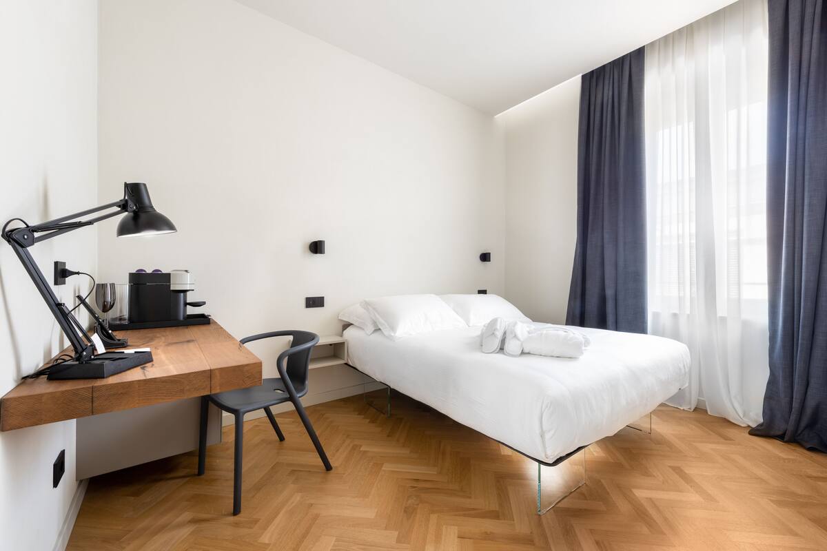 Quarto de hotel moderno com escrivaninha de madeira, cadeira preta, cama com roupa de cama branca e uma grande janela com cortinas.