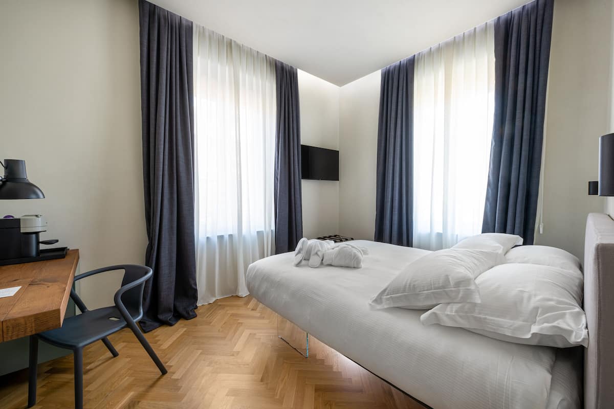 Quarto de hotel moderno com escrivaninha de madeira, uma cadeira, cama com roupa branca e televisão de tela plana fixada na parede.