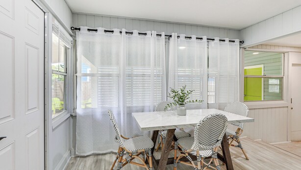 A dining area with a white table and four patterned chairs, a sheer curtain, and a view of a green door and a window with blinds. - Idyllic Sarasota Home w/ Yard: 2 Mi to Beach! (Sarasota)