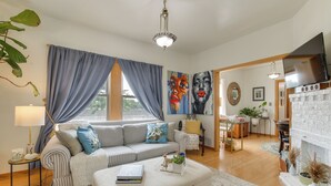 A living room with a sofa, ottoman, and a table with a book. There is a window with curtains, a painting on the wall, and a television on a stand. - Pet Friendly: 8 Mi to Chicago - Fenced Backyard! (Oak Park)