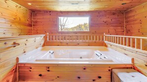 A wooden cabin room with a large bathtub, wooden walls, and a window with a view of trees. - Cozy Sevierville Cabin w/ Hot Tub, Mountain Views! (Sevierville)