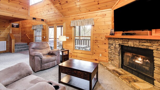 A cozy cabin interior with a stone fireplace, wooden walls, a leather sofa, and a television. - Cozy Sevierville Cabin w/ Hot Tub, Mountain Views! (Sevierville)