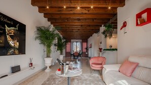 A modern living room with a wooden ceiling, a white sofa, a pink armchair, and a dining area in the background.