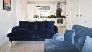 Un salon moderne avec un canapé bleu foncé et un pouf assorti. On aperçoit une cuisine à l’arrière-plan, avec des armoires blanches et des appareils électroménagers en acier inoxydable.