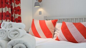Un lit bien fait, avec une literie blanche et des oreillers à rayures rouges et blanches, une serviette blanche enroulée et une lampe fixée au mur.