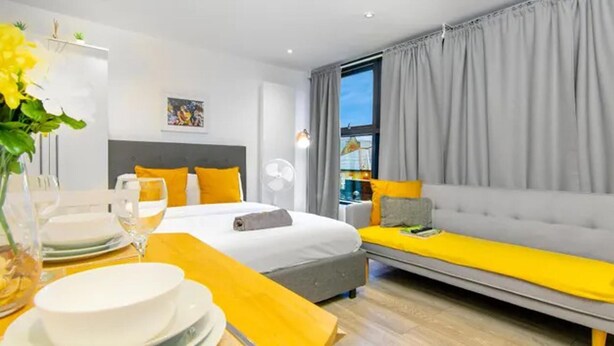 Moderni hotellihuone, jossa on sänky, sohva, ruokapöytä astioineen ja laseineen sekä suuri ikkuna, jossa on verhot.