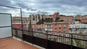 A balcony with a tiled floor and a metal railing, overlooking a cityscape with various buildings and a cloudy sky.