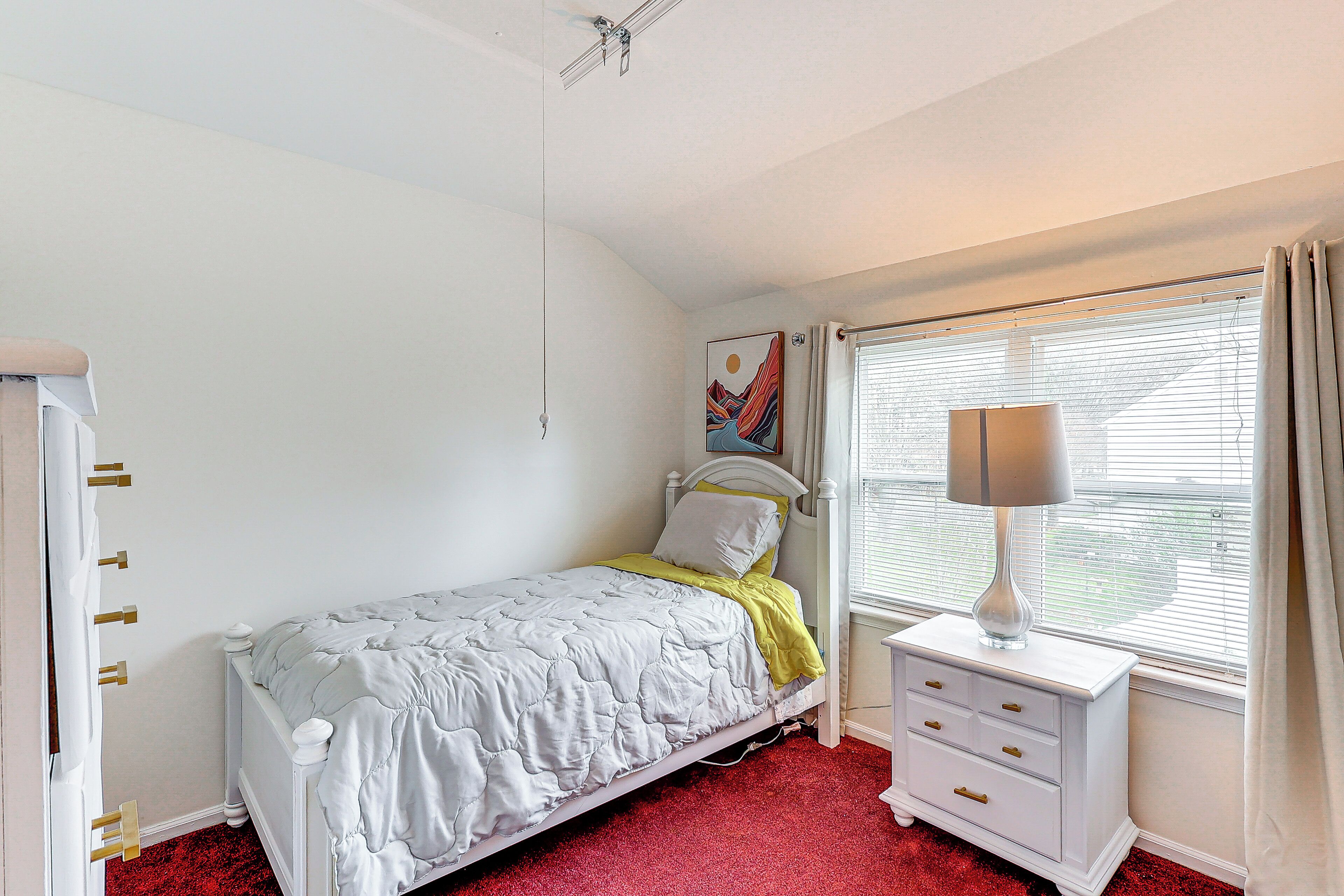 Une chambre à coucher comprenant un lit, une table de chevet, une lampe, une fenêtre avec des rideaux et un tableau mural.