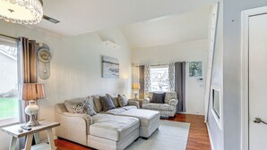 A living room with a sectional sofa, a small table, and a lamp. There is a window with curtains and a clock on the wall.
