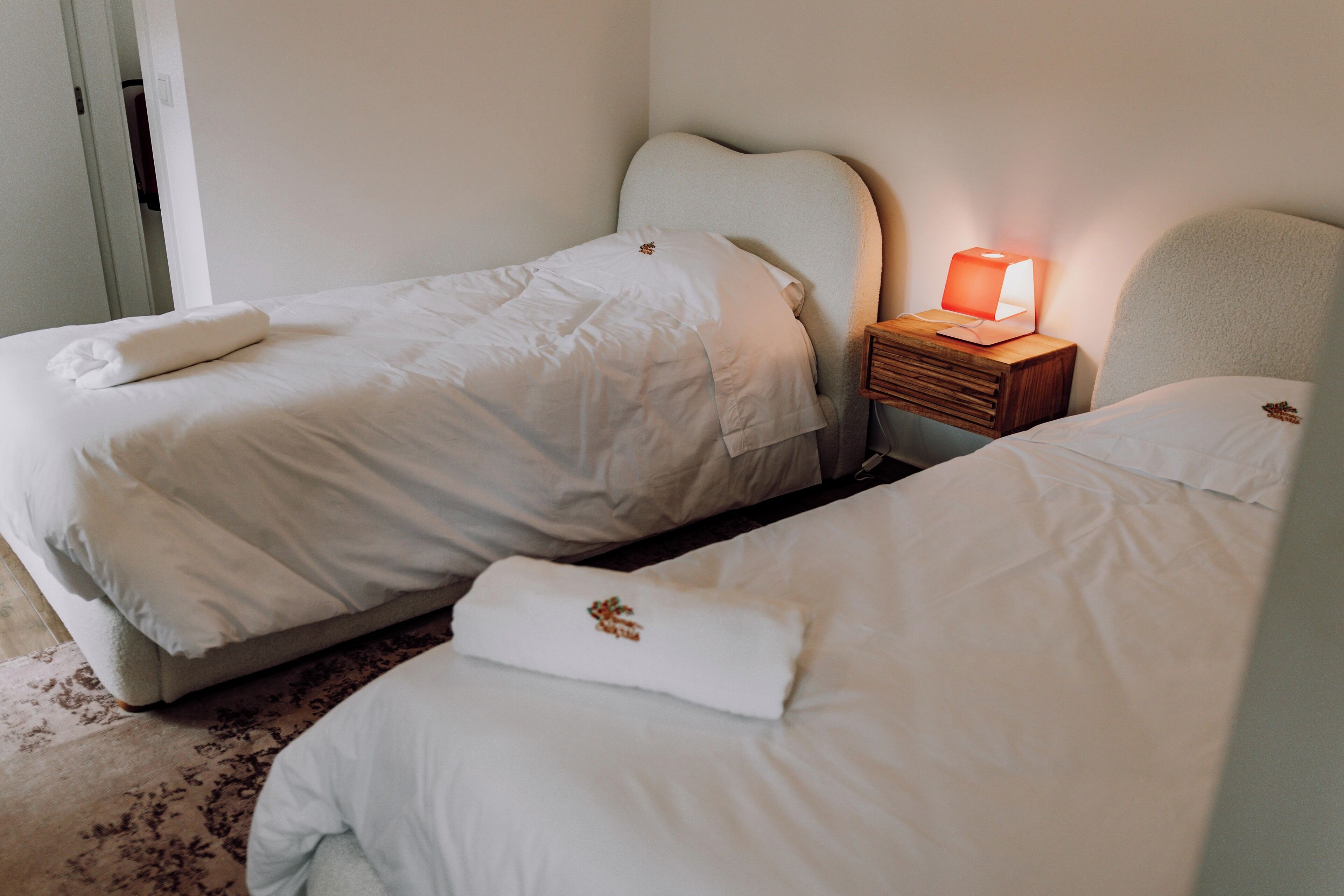 Habitación doble con dos camas, una mesita de noche con lámpara y una puerta que da a otra habitación.