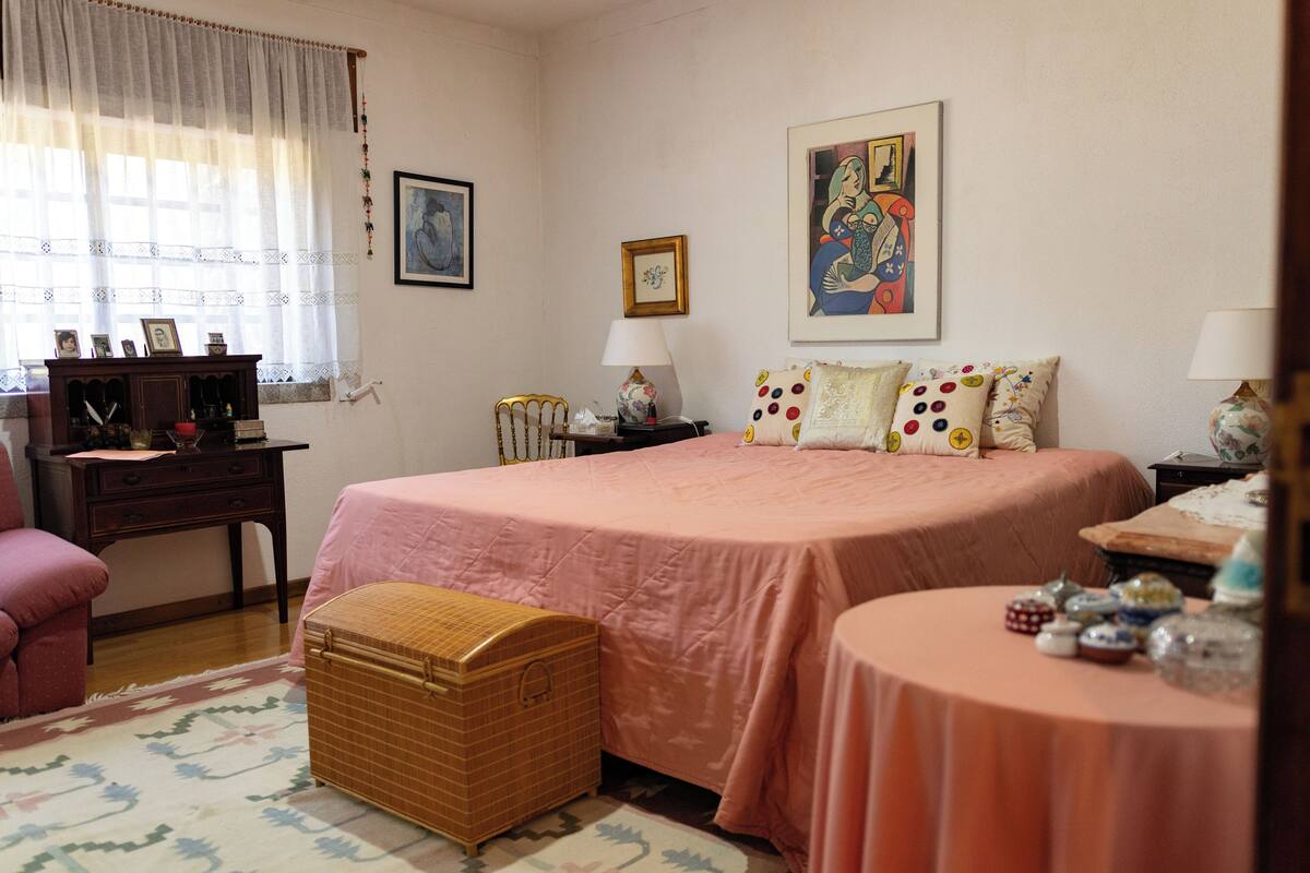 A bedroom with a large bed, a bedside table, a chair, a small table, and a chest of drawers.